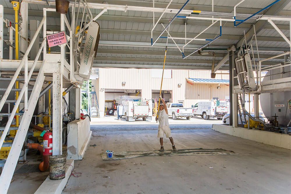 A person uses a pole to adjust equipment in a concrete industrial area. Multiple vehicles are parked outside.