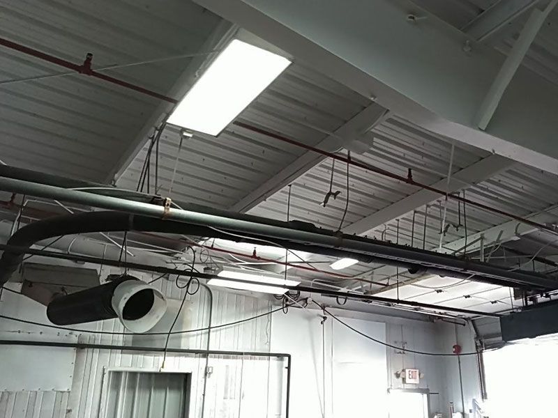 Industrial ceiling with white painted beams, exposed pipes, ventilation, and fluorescent lights.