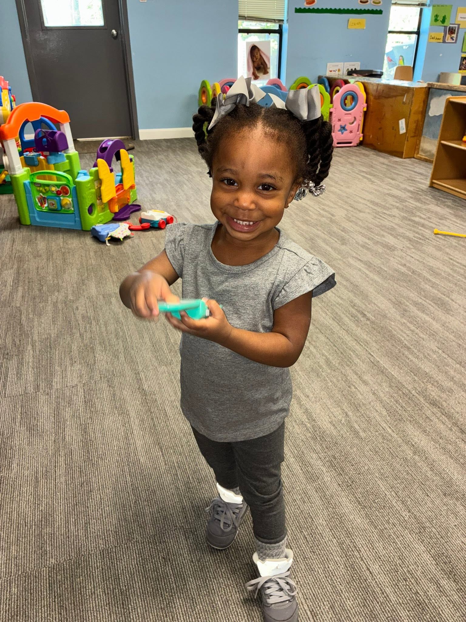 Smiling child standing indoors, holding a small toy in a playroom with colorful toys and carpet.