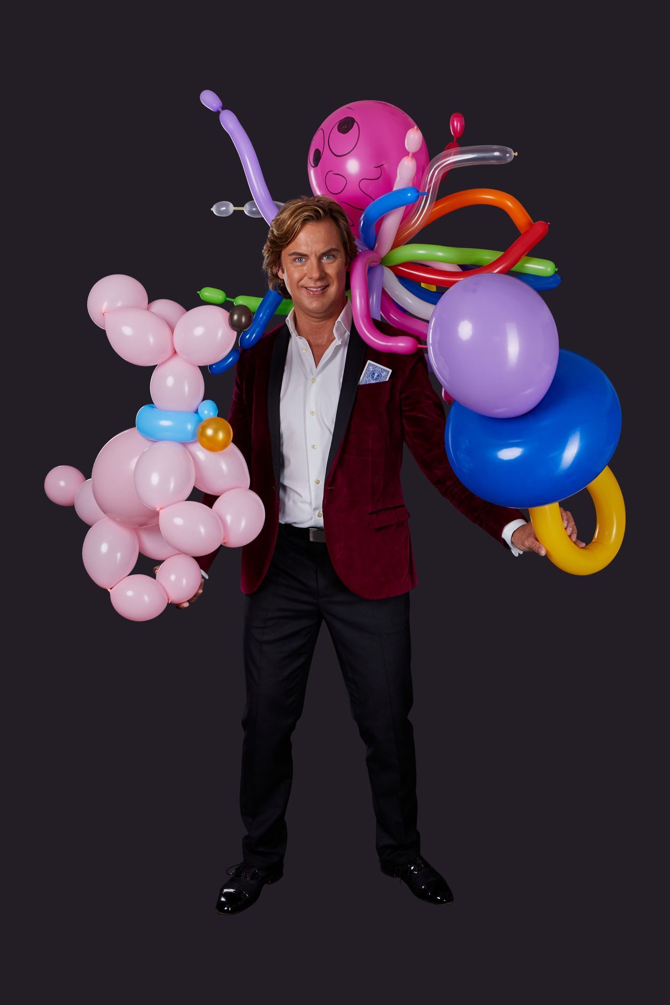 Man in rode blazer met ballonfiguren en kleurrijke ballonnen.