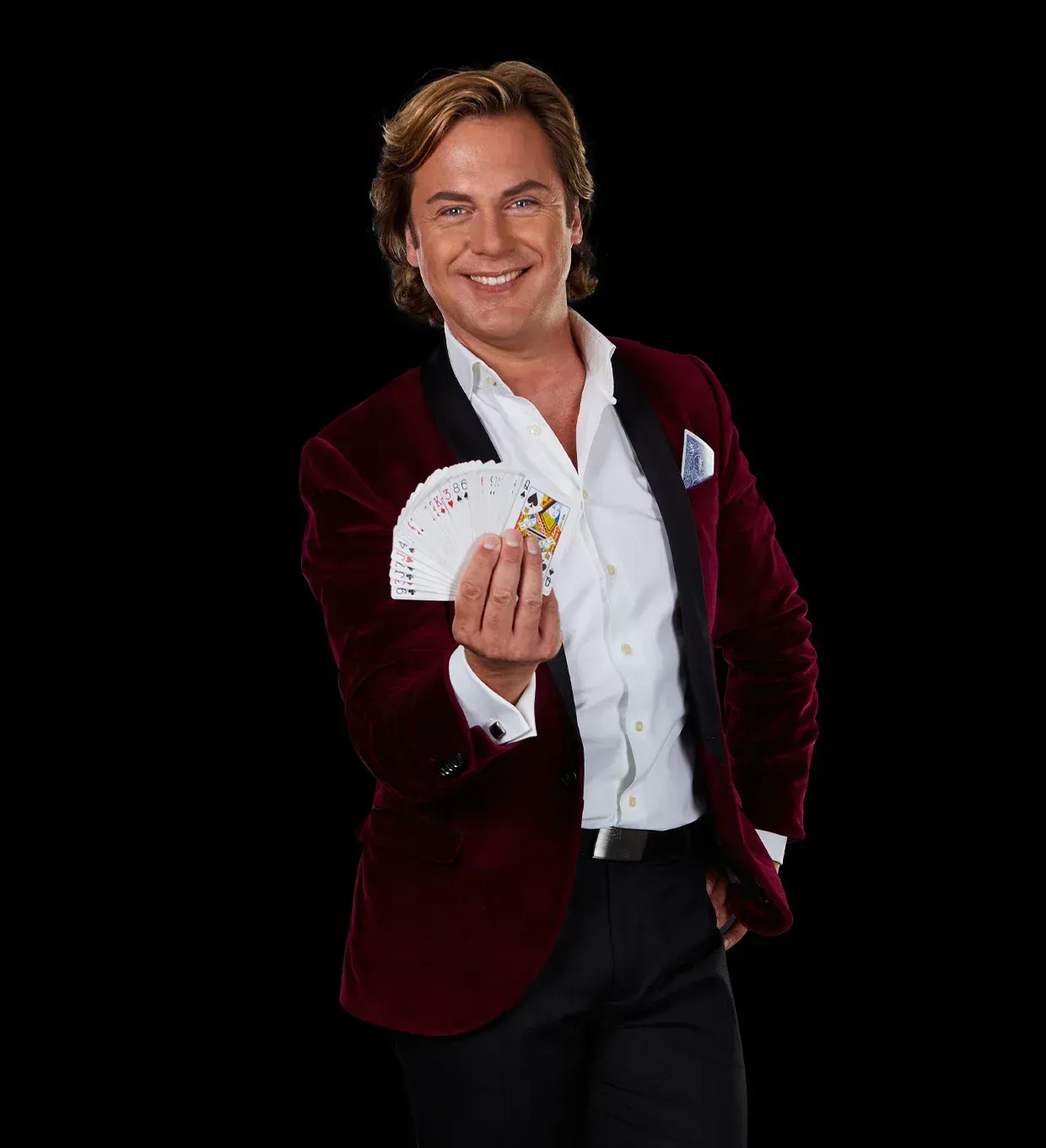 Een goochelaar in een bordeauxrode jas met een uitgevouwen pak speelkaarten. Hij lacht en staat voor een zwarte achtergrond.