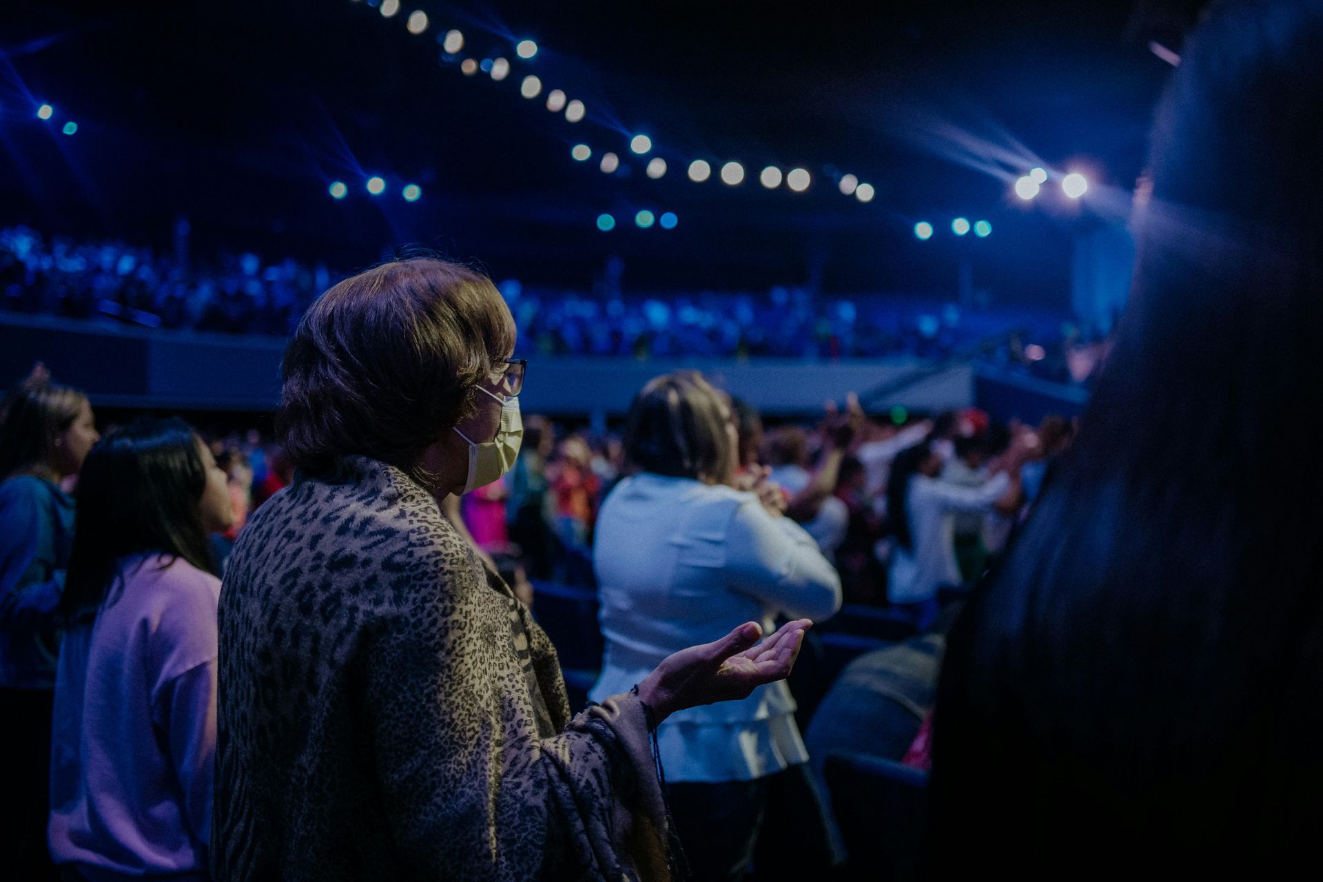 People raising hands in a crowded auditorium, likely a worship service. Blue lighting, spotlights.