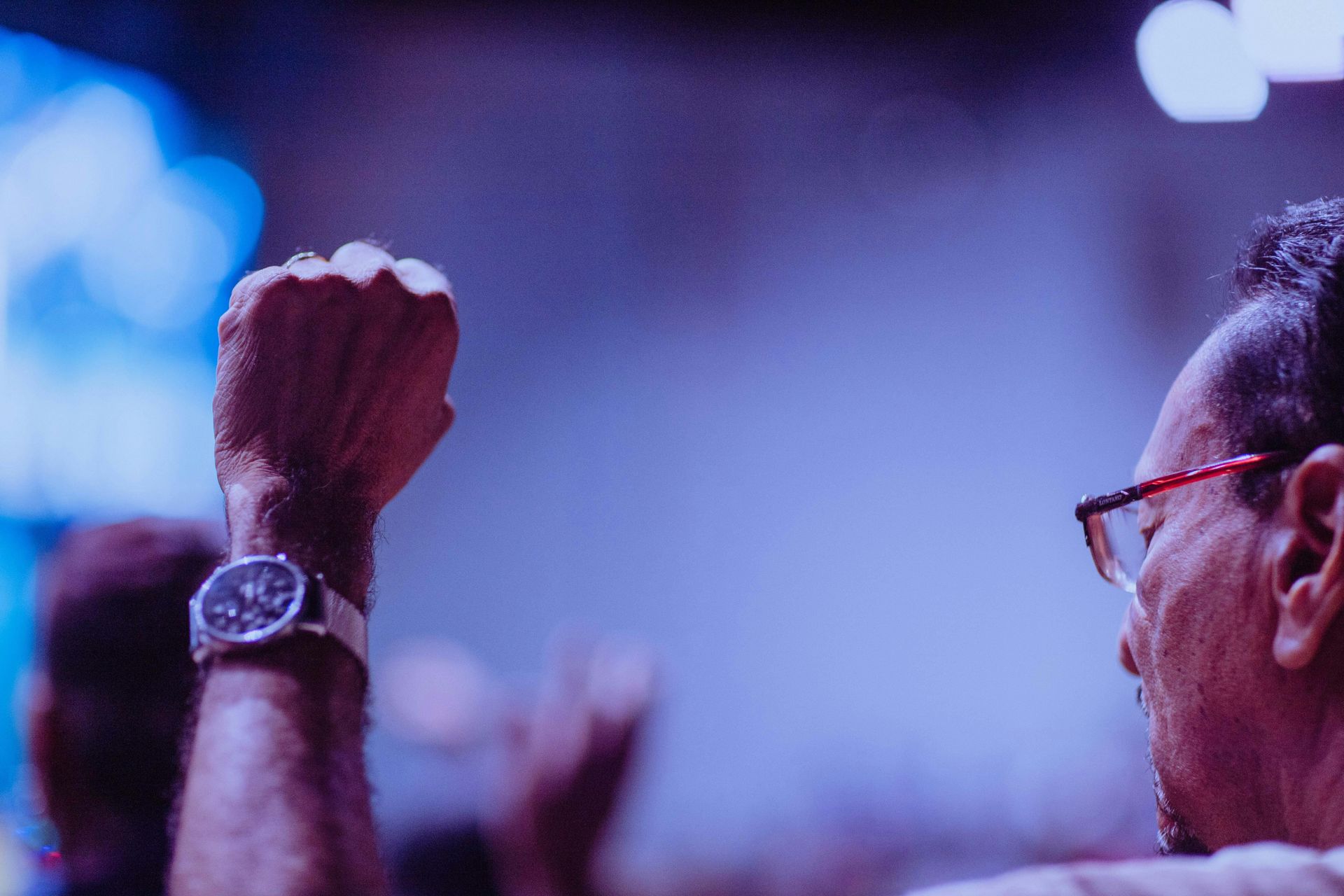 Person with glasses raises a fist in a crowd, watch visible. Blue-toned lighting.