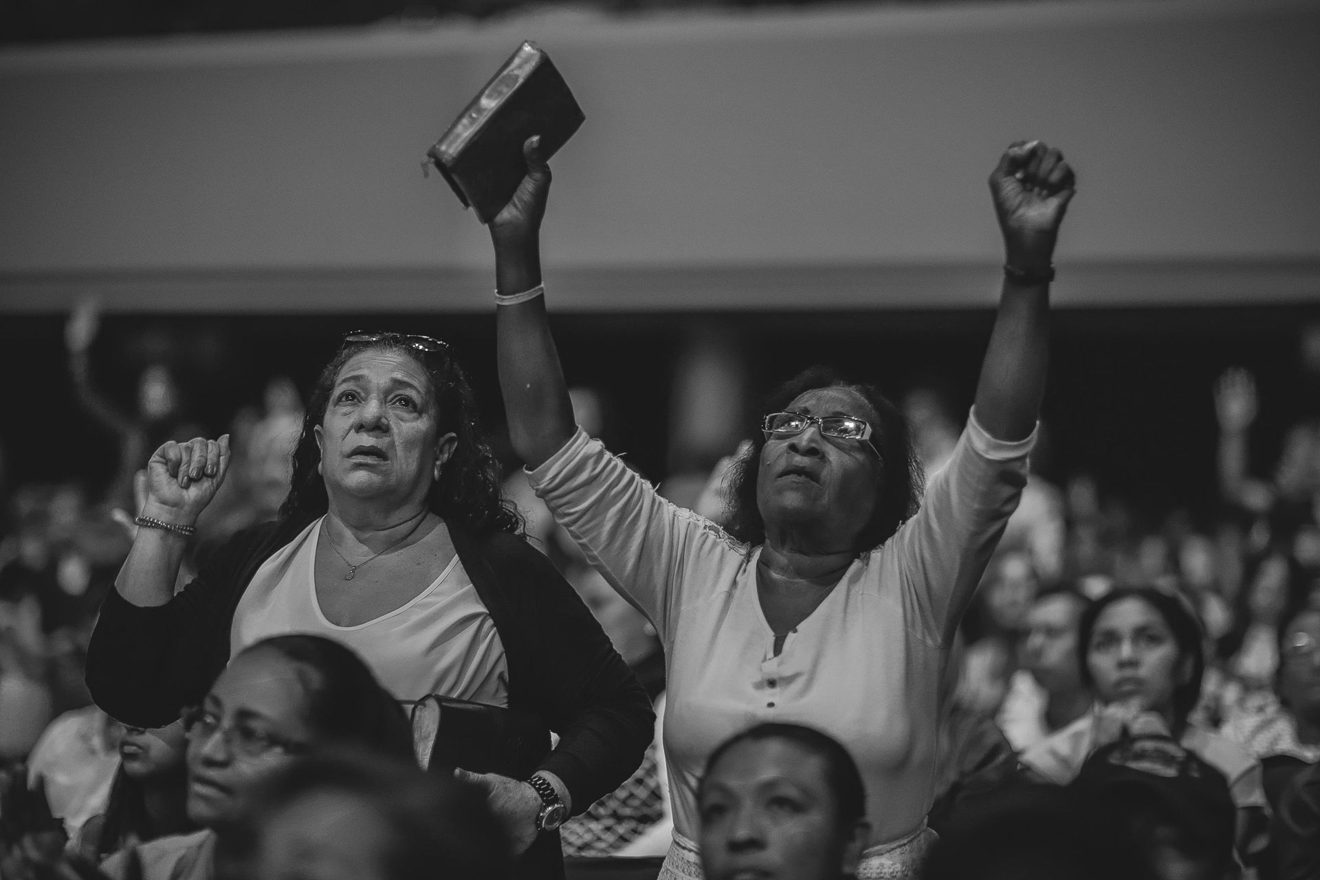 People in a crowded room with raised arms, one holding a book. Black and white photo.