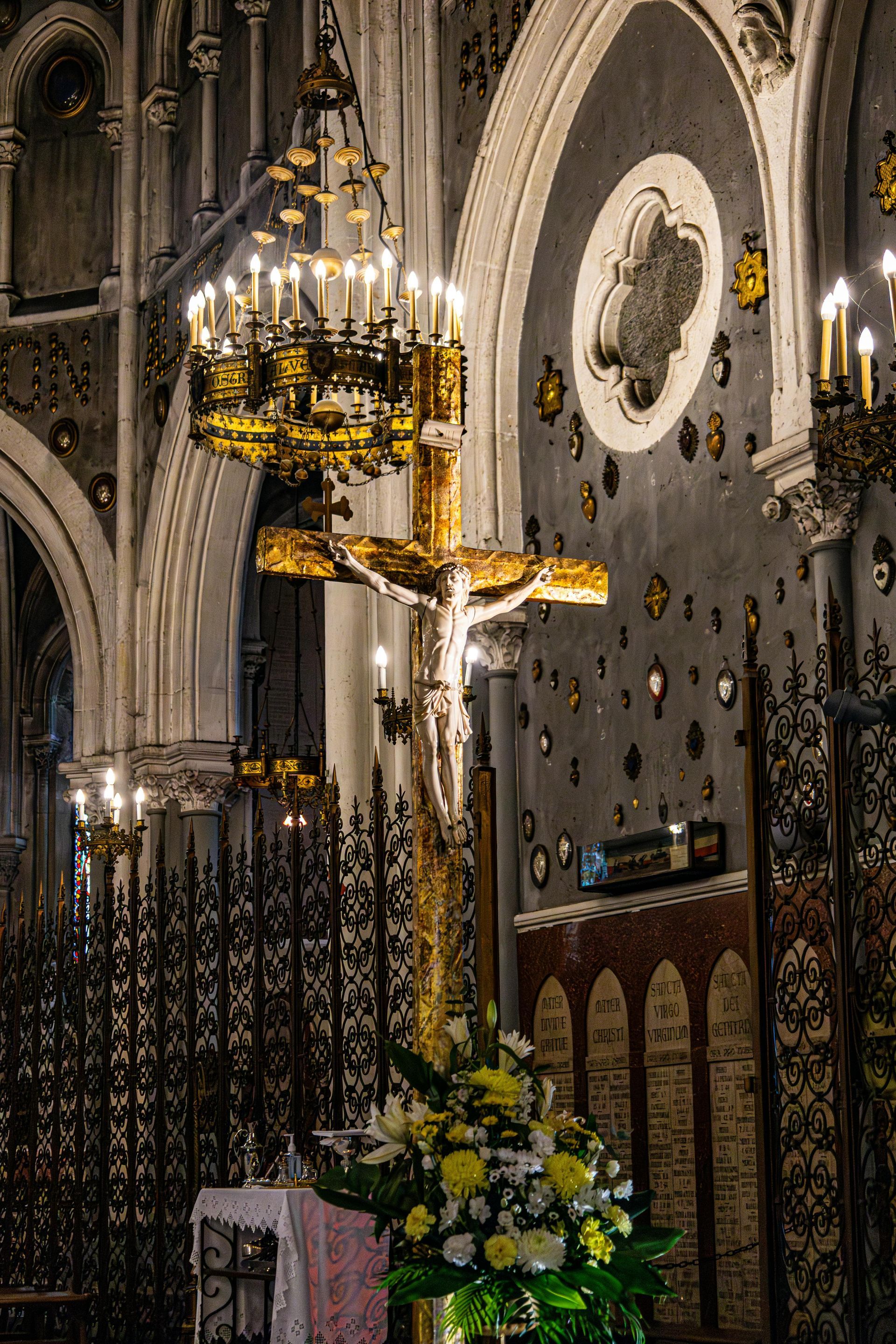 Gilded crucifix in a church, flanked by candles and flowers. Dark, ornate interior with arched ceilings.