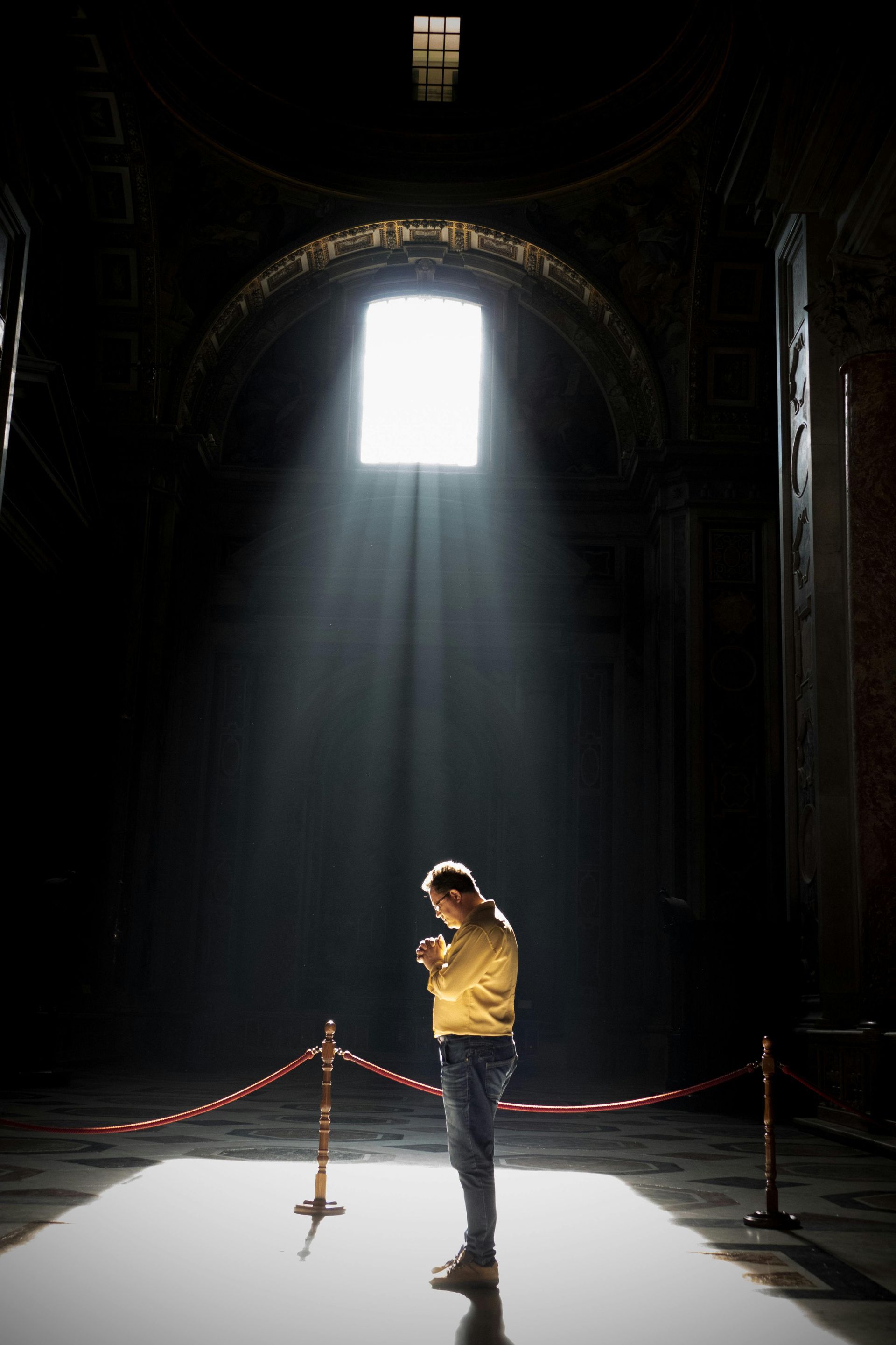 Person standing in dark, arched room with a bright light shining from a window, golden-yellow shirt and blue jeans.