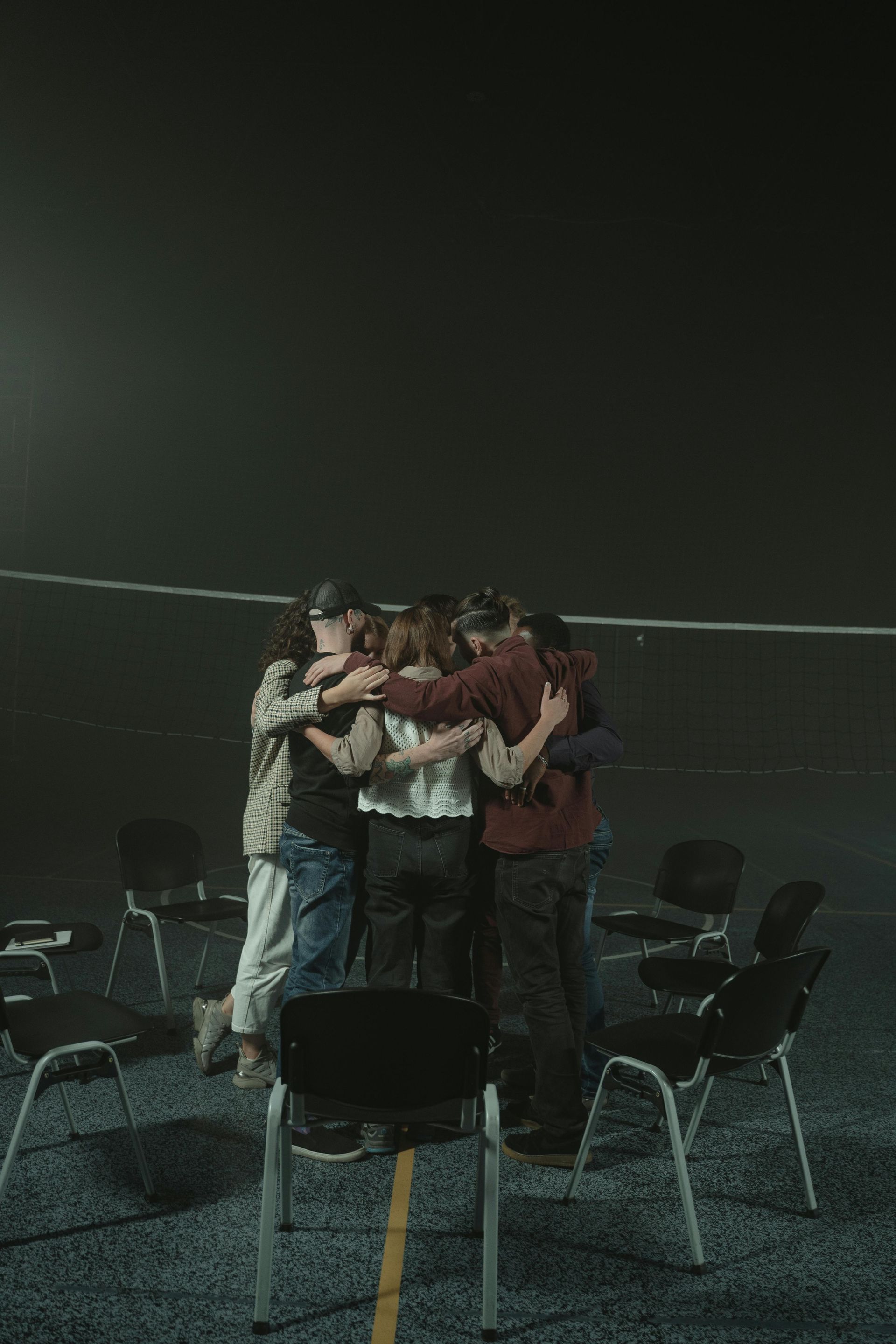 A group of people embrace in a circle. They stand in a dark room with chairs around them.