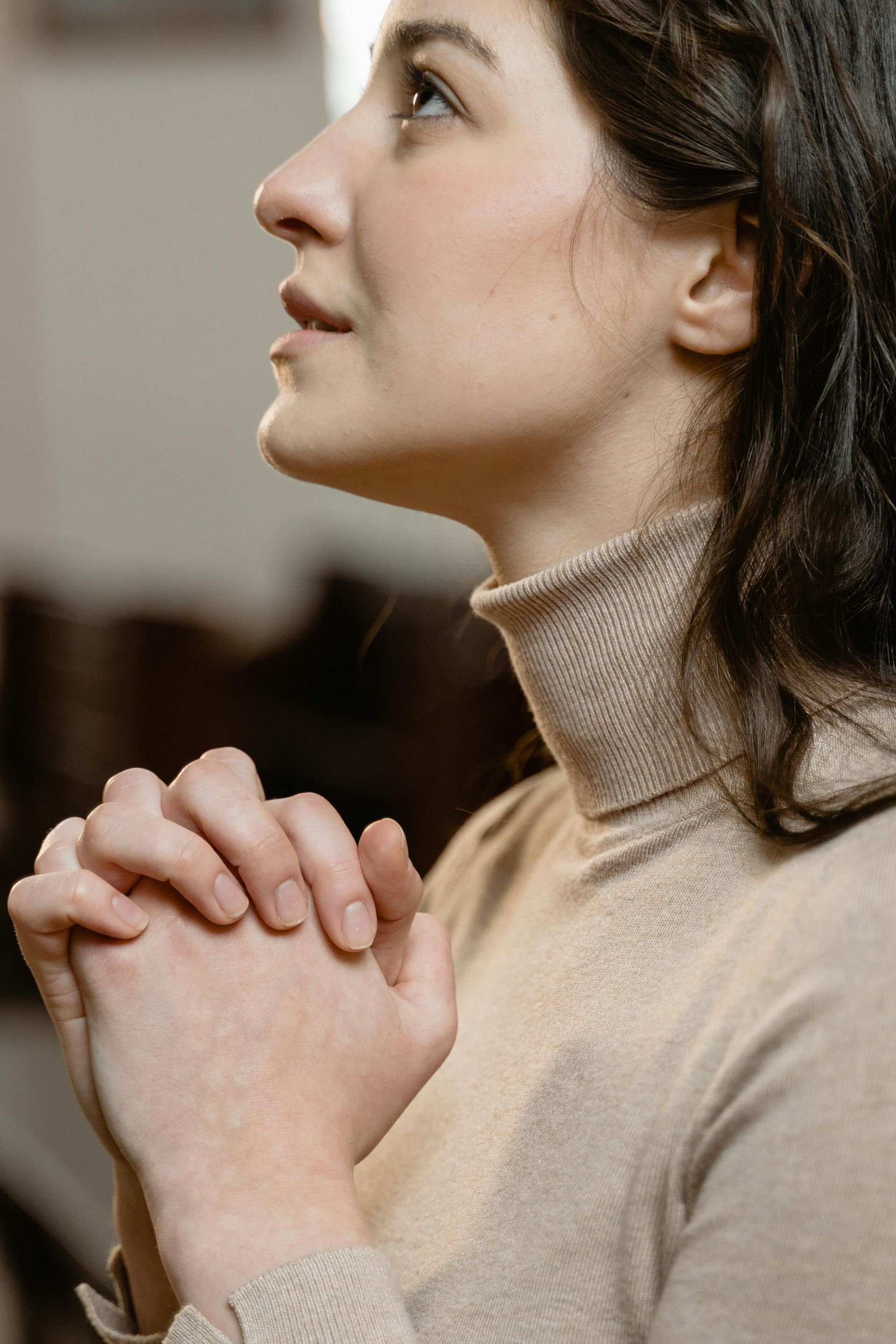 Woman clasps hands, gazes upward in prayer, wearing a tan turtleneck. Soft lighting, blurred background.