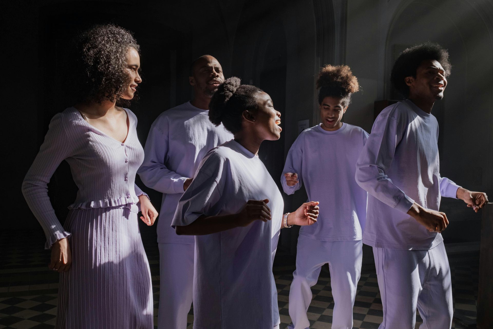 Choir in white outfits sing, lit by sunlight in a dim room.