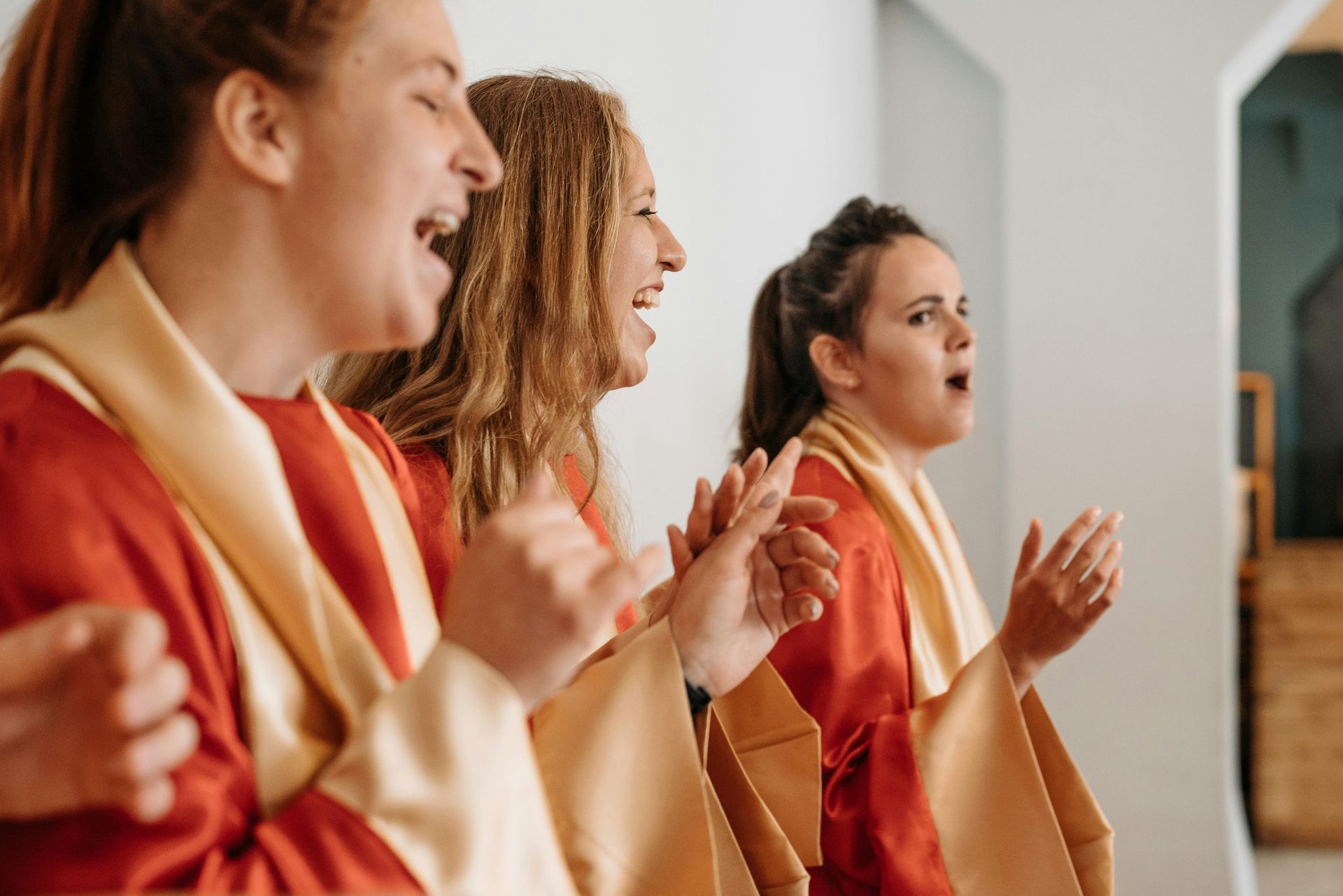 Three people in red and gold robes singing with open mouths and clapping hands.