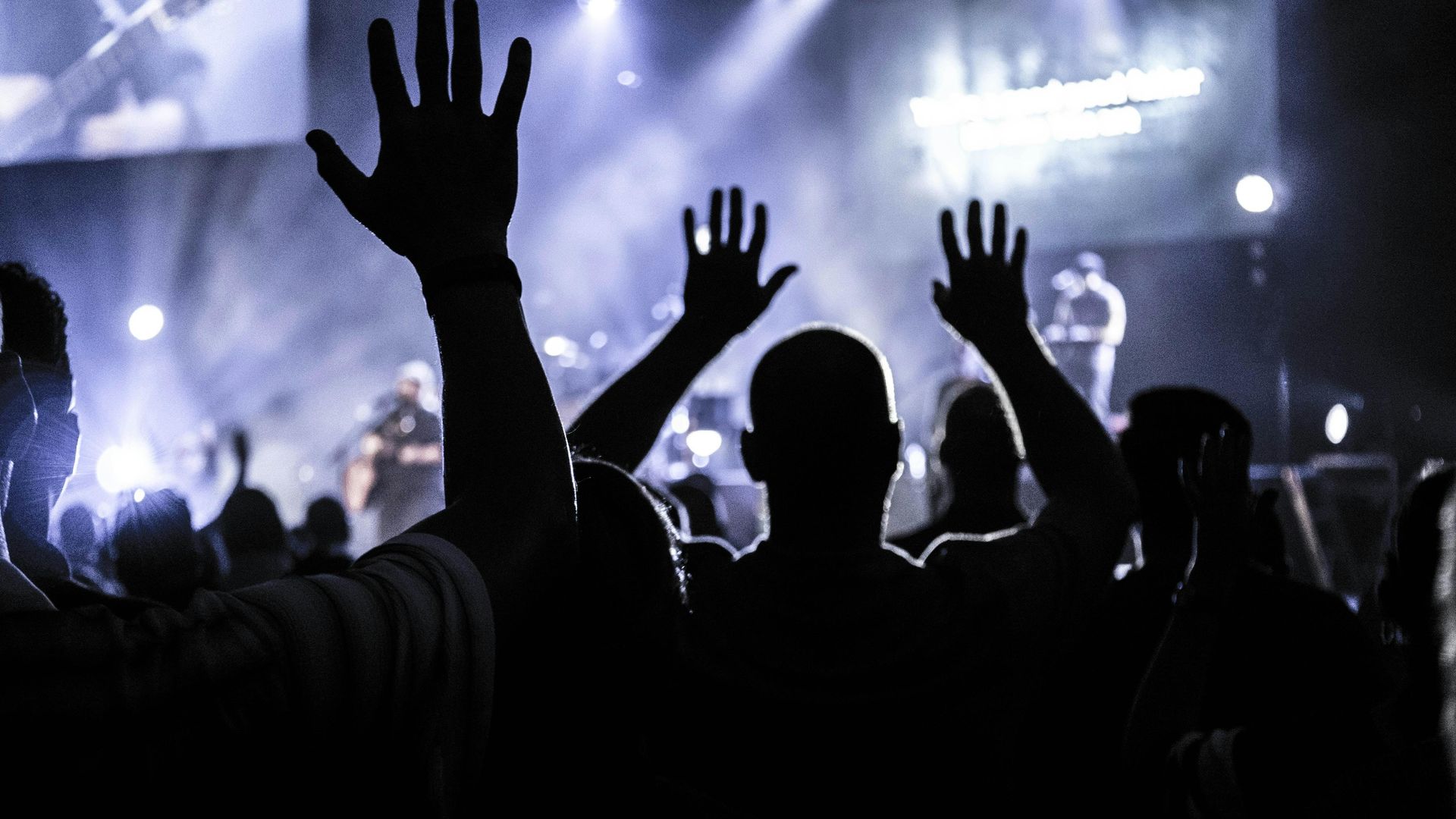 People with hands raised at a concert, silhouetted against a stage with lights.