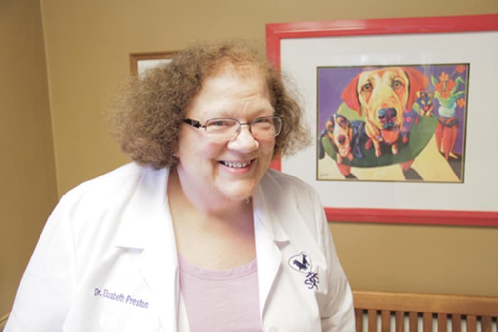 Woman in a white coat and glasses smiles, a colorful dog portrait behind her.