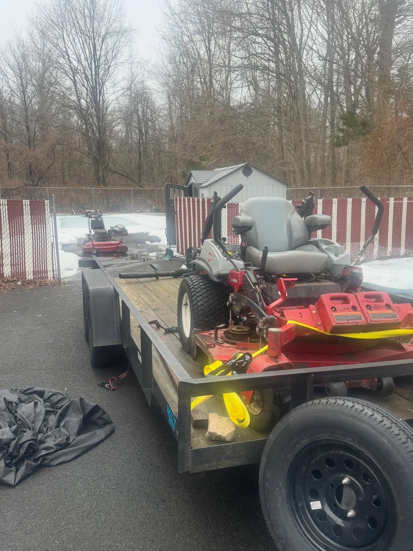 Small utility trailer carrying a red lawn tractor on a wet road with a striped barrier and trees behind it
