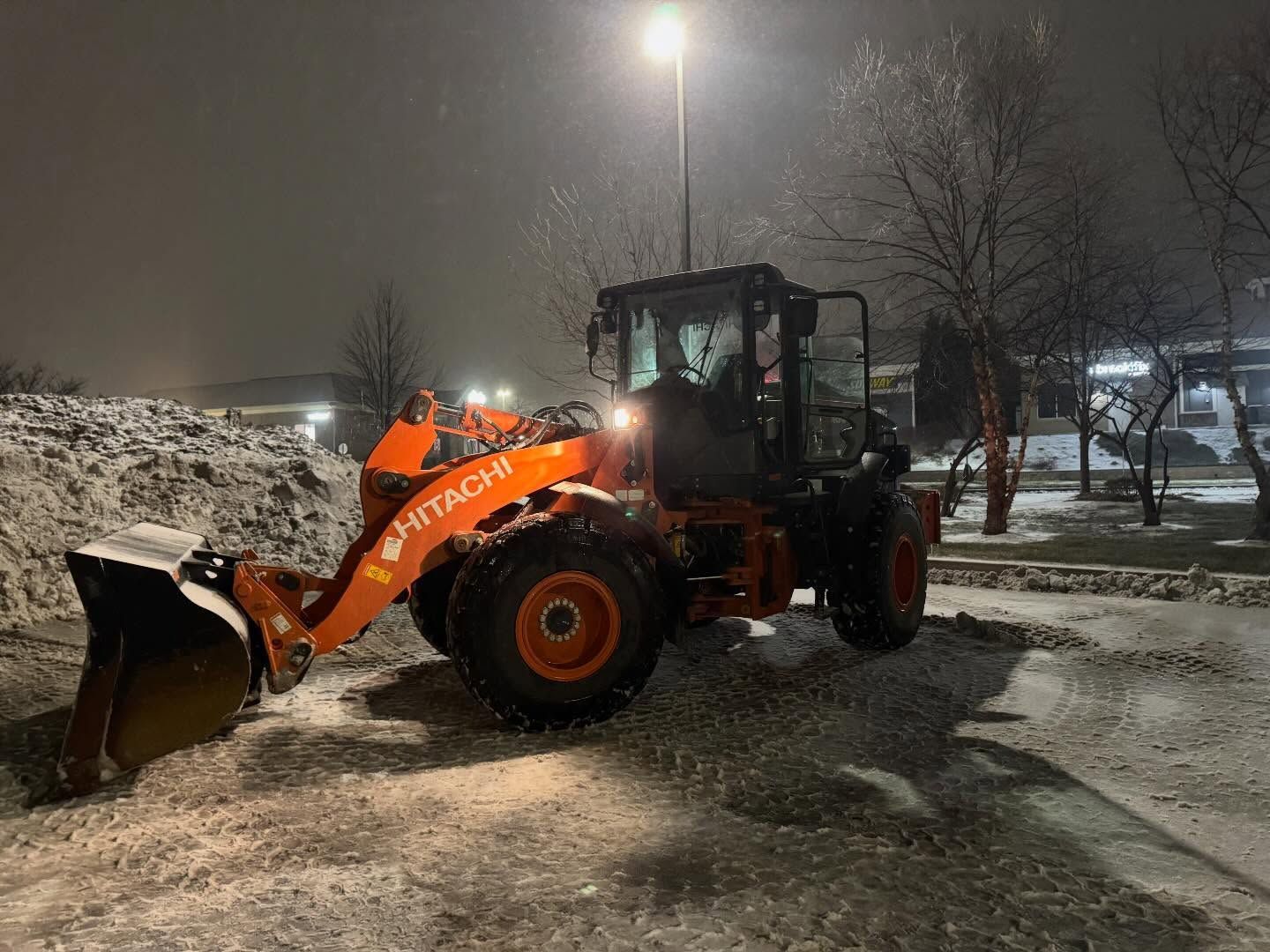 Orange snowplow clearing snow in a parking lot at night under a streetlight.