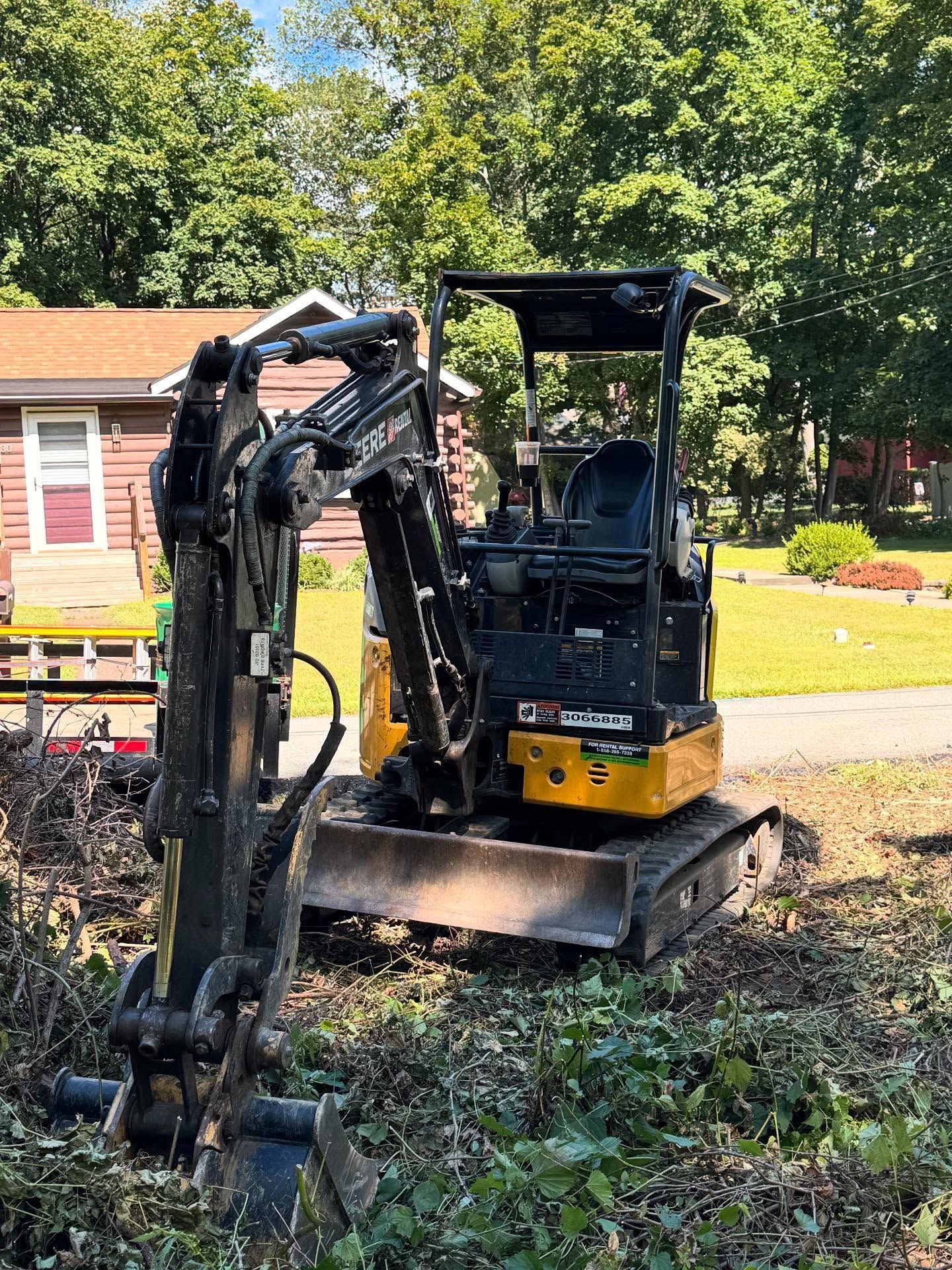 Compact excavator clearing brush in a yard near a small house with trees in the background