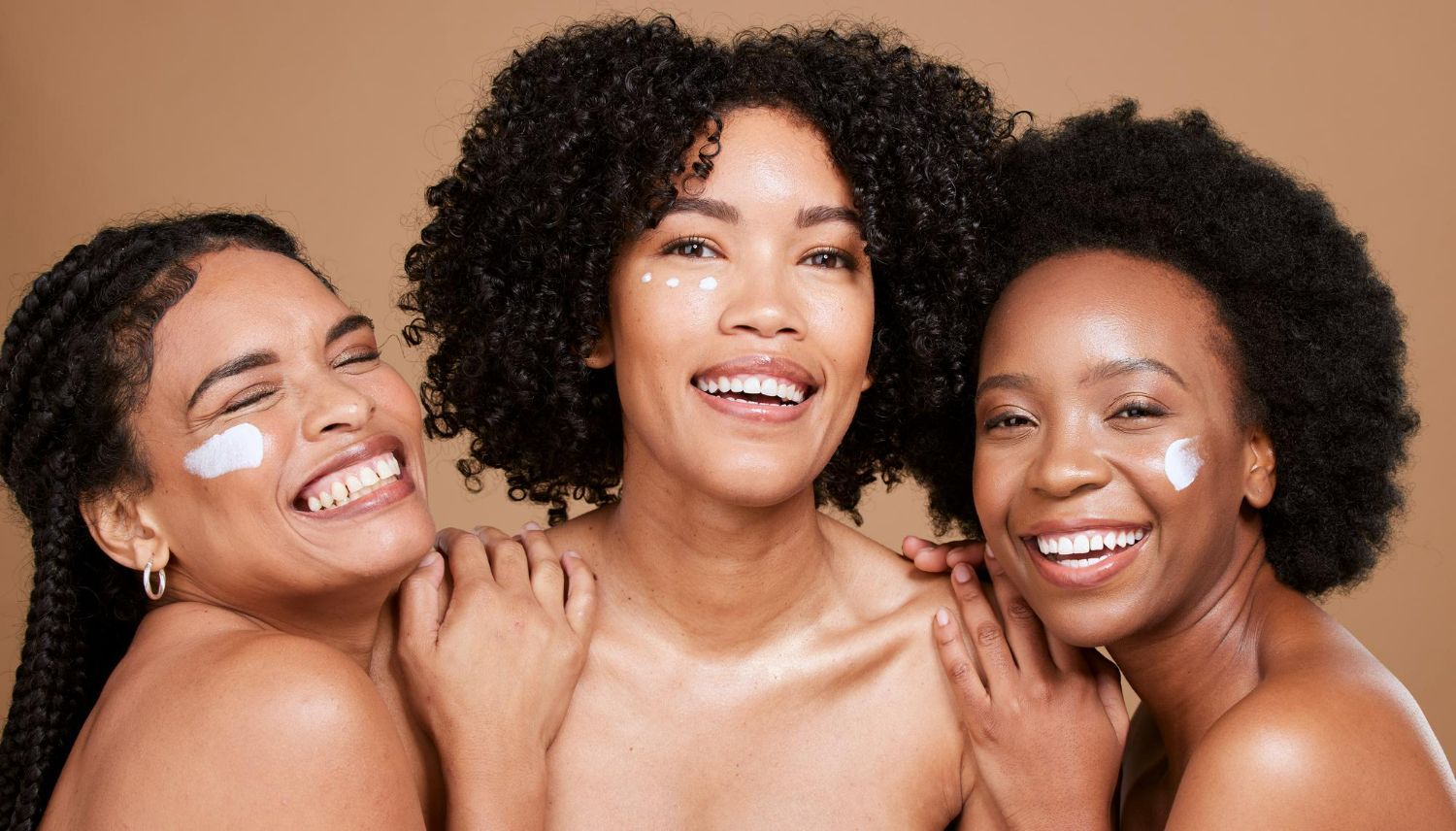 Three smiling people with textured hair and skin cream on their cheeks posing together against a plain tan background.