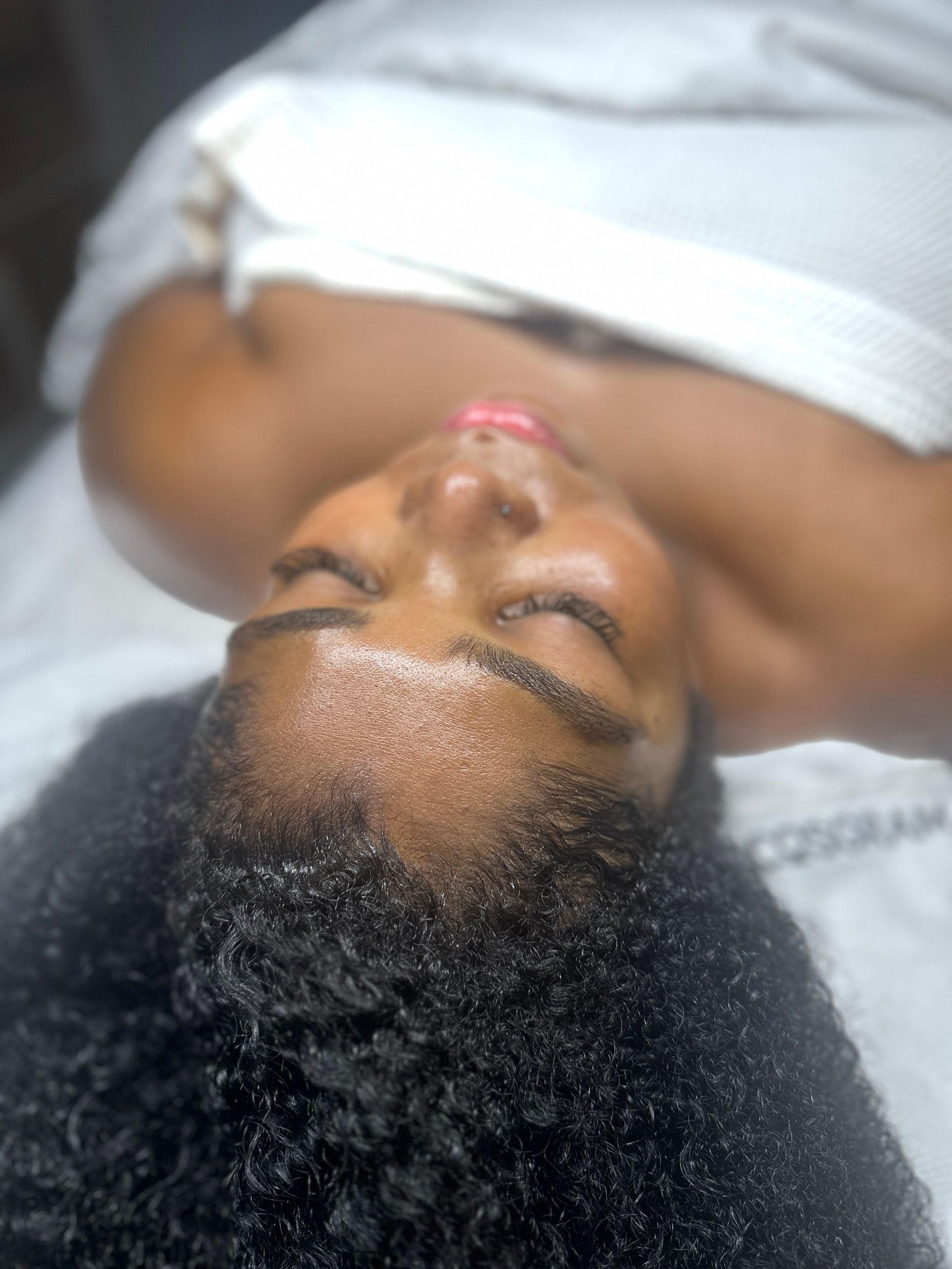 Person lying on a bed with eyes closed, wearing a white top, with curly hair spread out on the pillow.