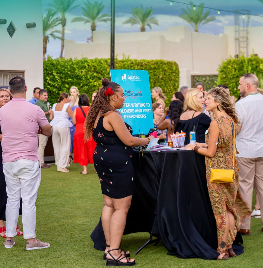 People mingle at an outdoor event with a table. Sign says