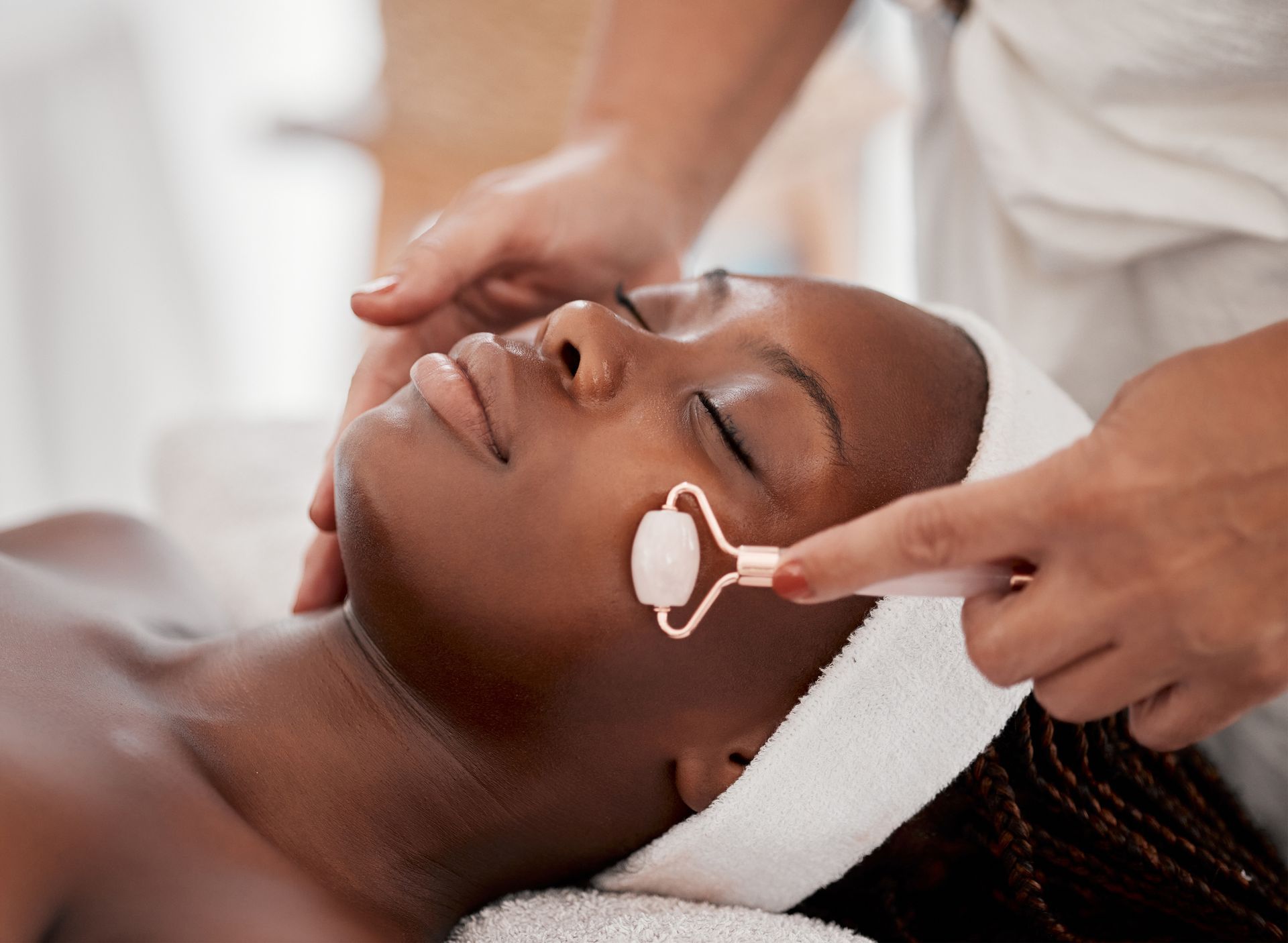 Facial treatment on a reclining client with a white headband and jade roller on the cheek