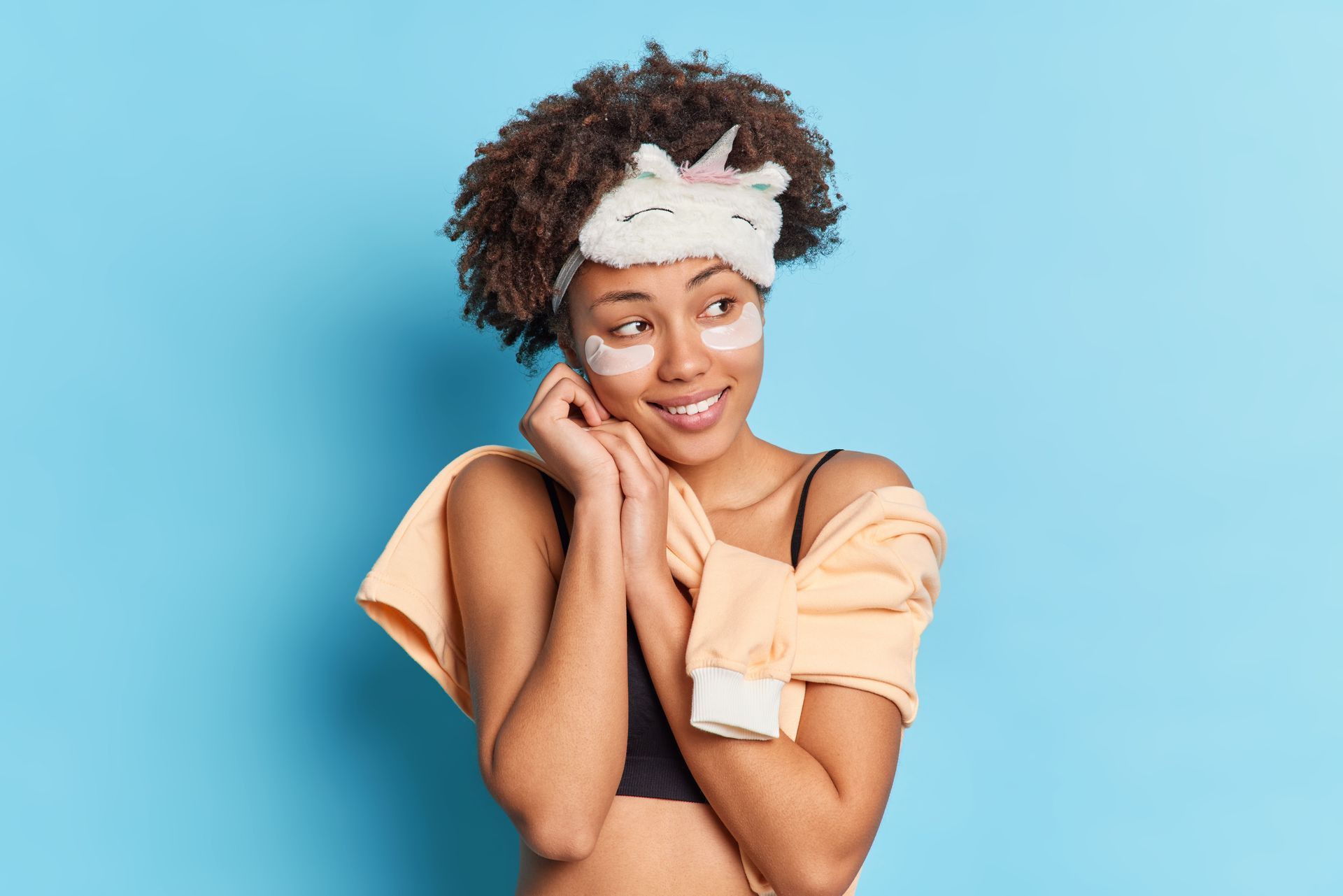 Woman smiling with hands on cheeks against a blue background, wearing a white headband and peach top
