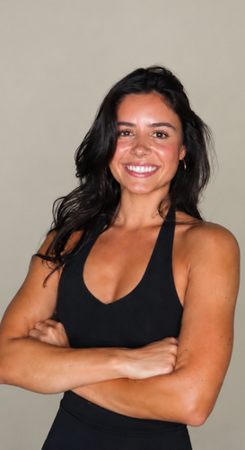 A person in black workout gear with tattoos on their arms and ribs stands with palms pressed together, smiling.