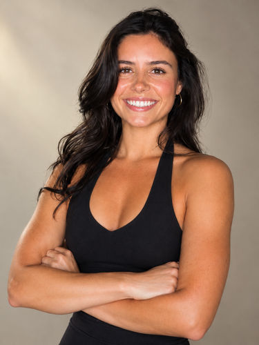 A person in black workout gear with tattoos on their arms and ribs stands with palms pressed together, smiling.