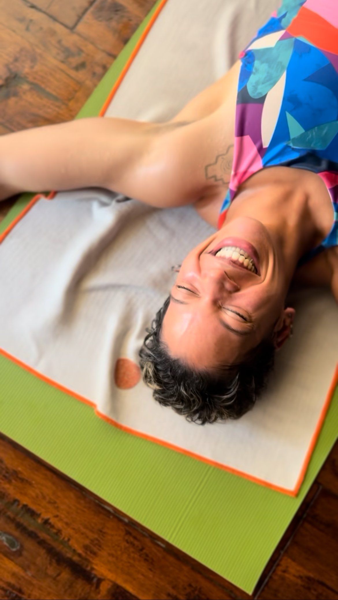 A smiling person lying on a yoga mat with a patterned towel, arms outstretched, viewed from above on a wooden floor.