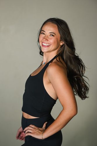A person with shoulder-length blonde hair smiles while wearing a black tank top against a plain background.