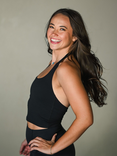 A person with shoulder-length blonde hair smiles while wearing a black tank top against a plain background.