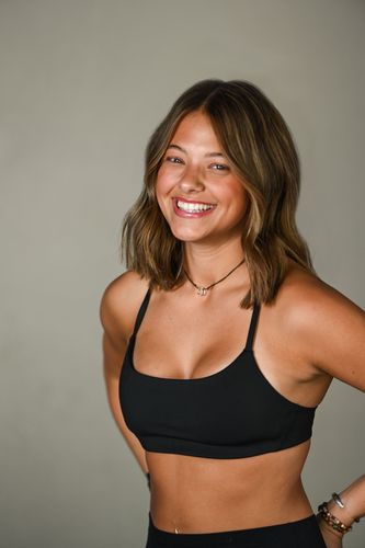 A smiling person with a beard, wearing a black t-shirt and dark shorts, reaching toward the camera against a gray wall.