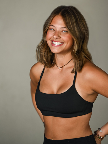 A smiling person with a beard, wearing a black t-shirt and dark shorts, reaching toward the camera against a gray wall.