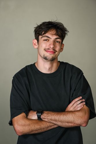 A smiling person wearing a black t-shirt makes a shaka hand gesture against a plain background.