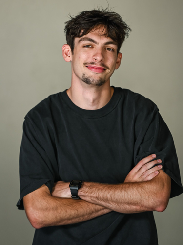 A smiling person wearing a black t-shirt makes a shaka hand gesture against a plain background.