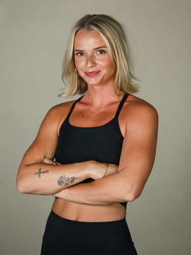 A smiling person in black athletic wear poses with hands on hips against a neutral background.