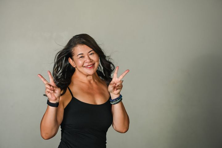 A smiling person wearing a black t-shirt makes a shaka hand gesture against a plain background.