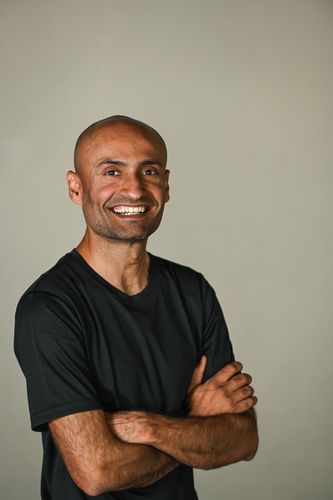 A smiling person wearing a black t-shirt makes a shaka hand gesture against a plain background.