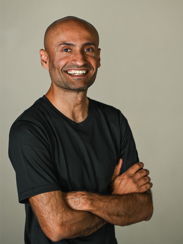 A smiling person wearing a black t-shirt makes a shaka hand gesture against a plain background.