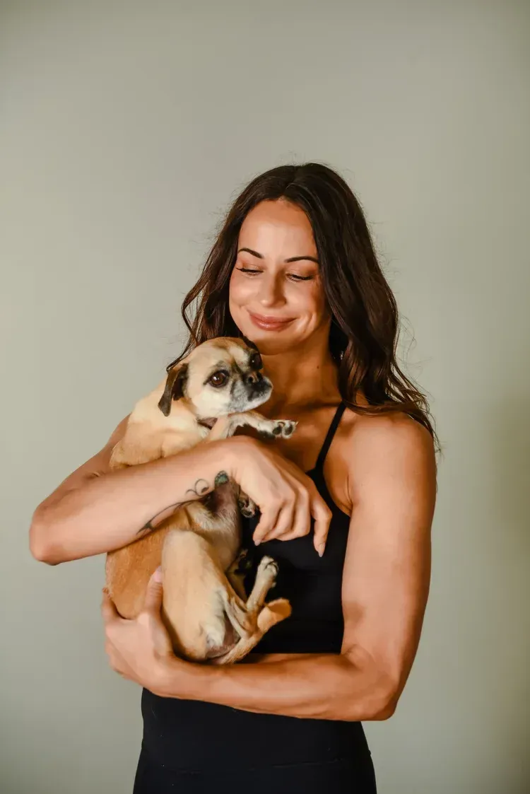 A person with long dark hair holds a small, light-brown dog close to their chest while smiling against a plain background.