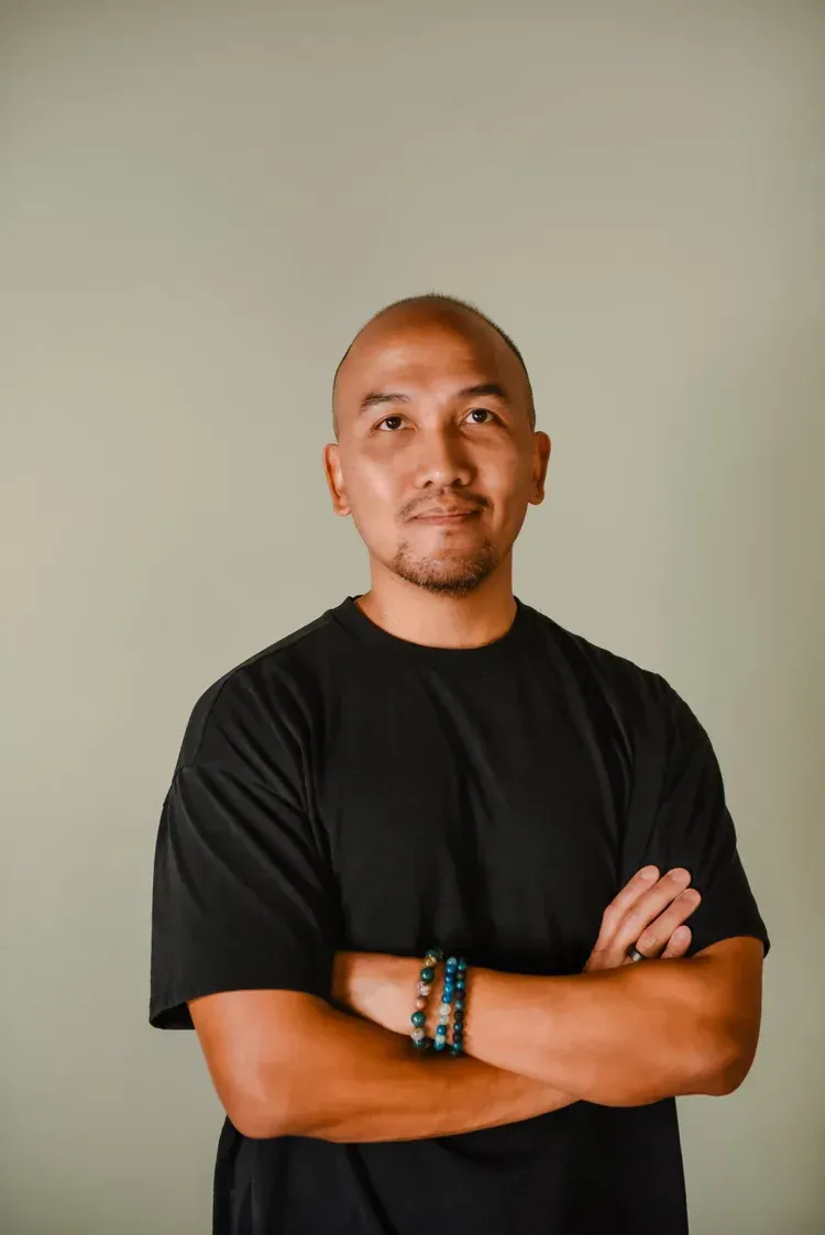 A person with a shaved head wearing a black t-shirt, smiling with arms crossed, set against a plain light background.