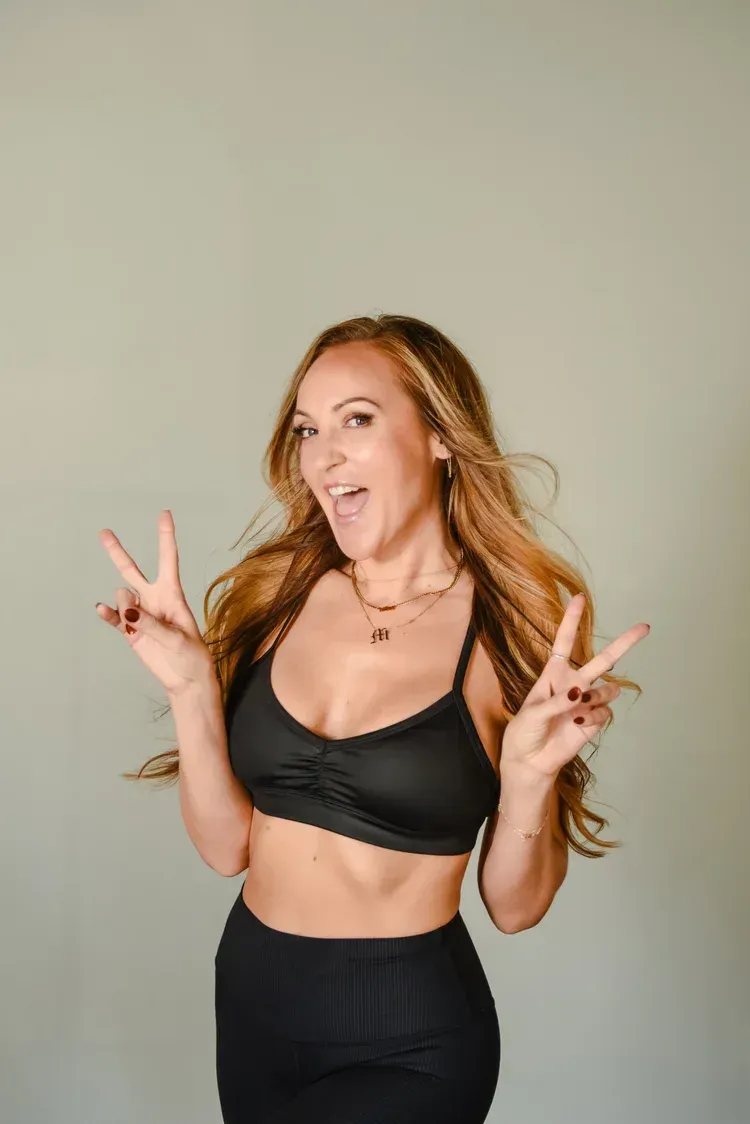 A person with long blonde hair smiles, wearing a black sports bra and leggings, holding up peace signs with both hands.