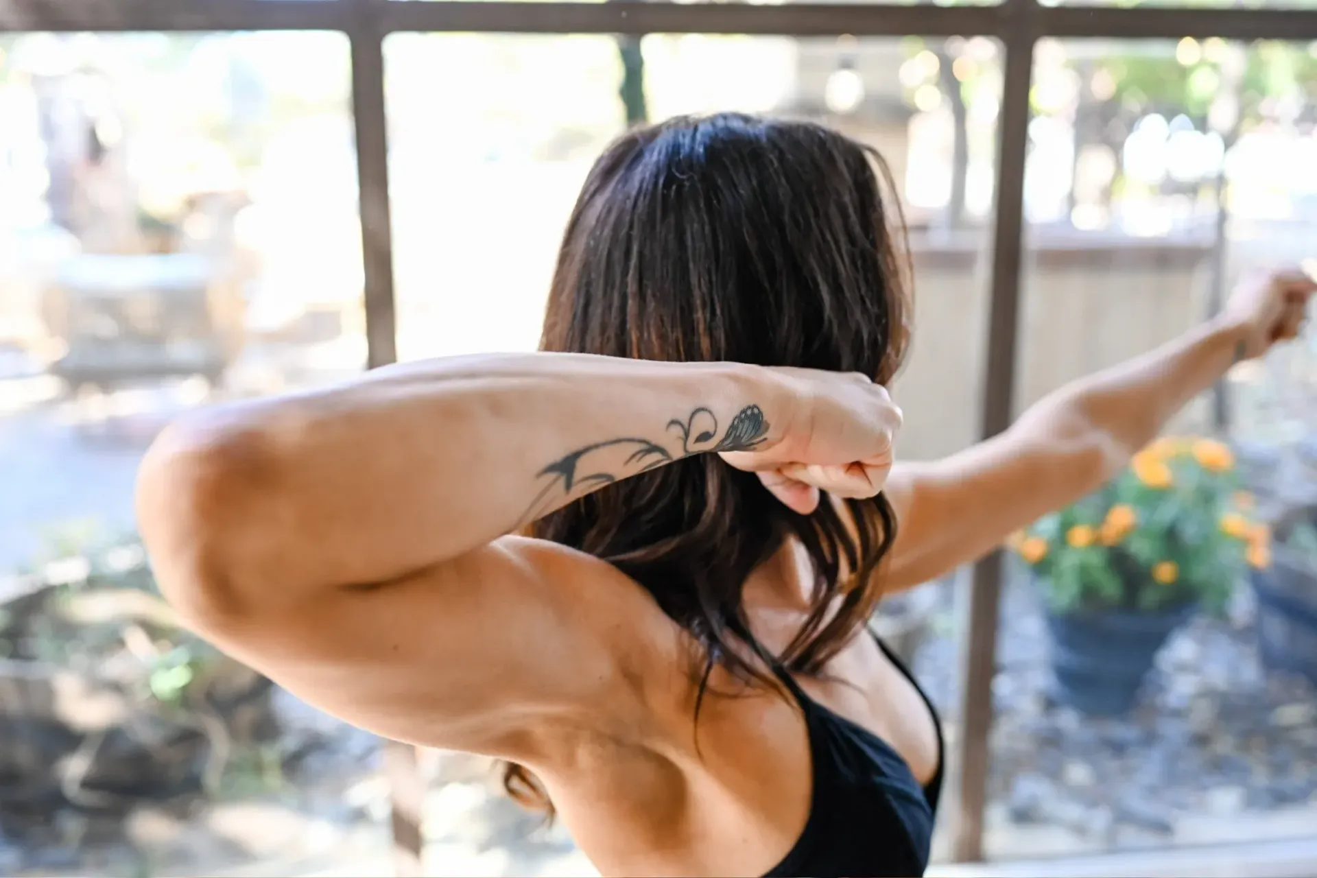 A person in a black sports bra flexing their arms and shoulders with a tattoo visible on their forearm.