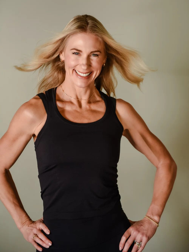 A person with shoulder-length blonde hair smiles while wearing a black tank top against a plain background.
