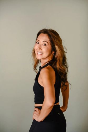 A smiling person with long hair wearing a black athletic top and leggings, posing with hands on hips against a plain wall.