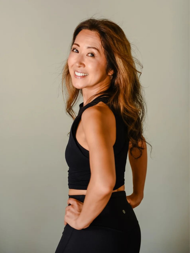 A smiling person with long hair wearing a black athletic top and leggings, posing with hands on hips against a plain wall.