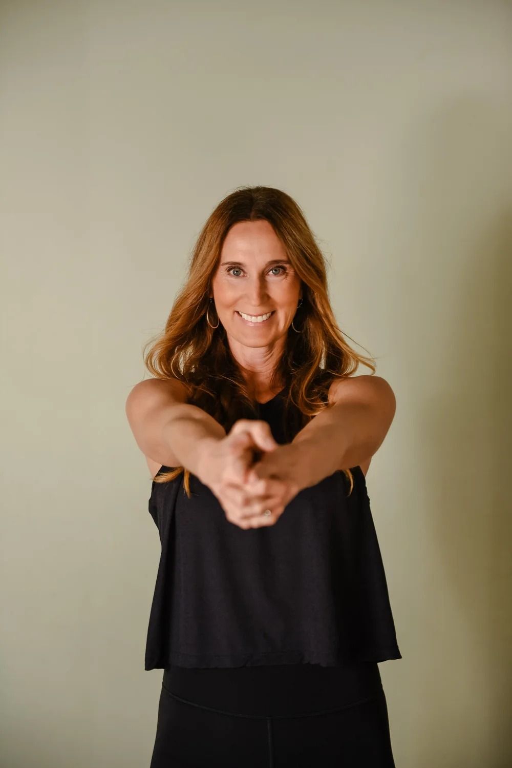 A person with long brown hair, wearing a black tank top, smiling while holding their hands together in front of them.