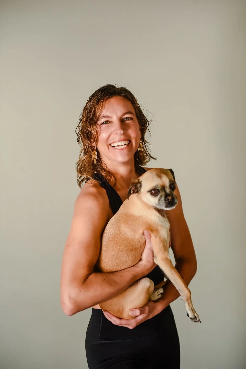 A smiling person with curly brown hair holding a tan, small dog against a light grey background.