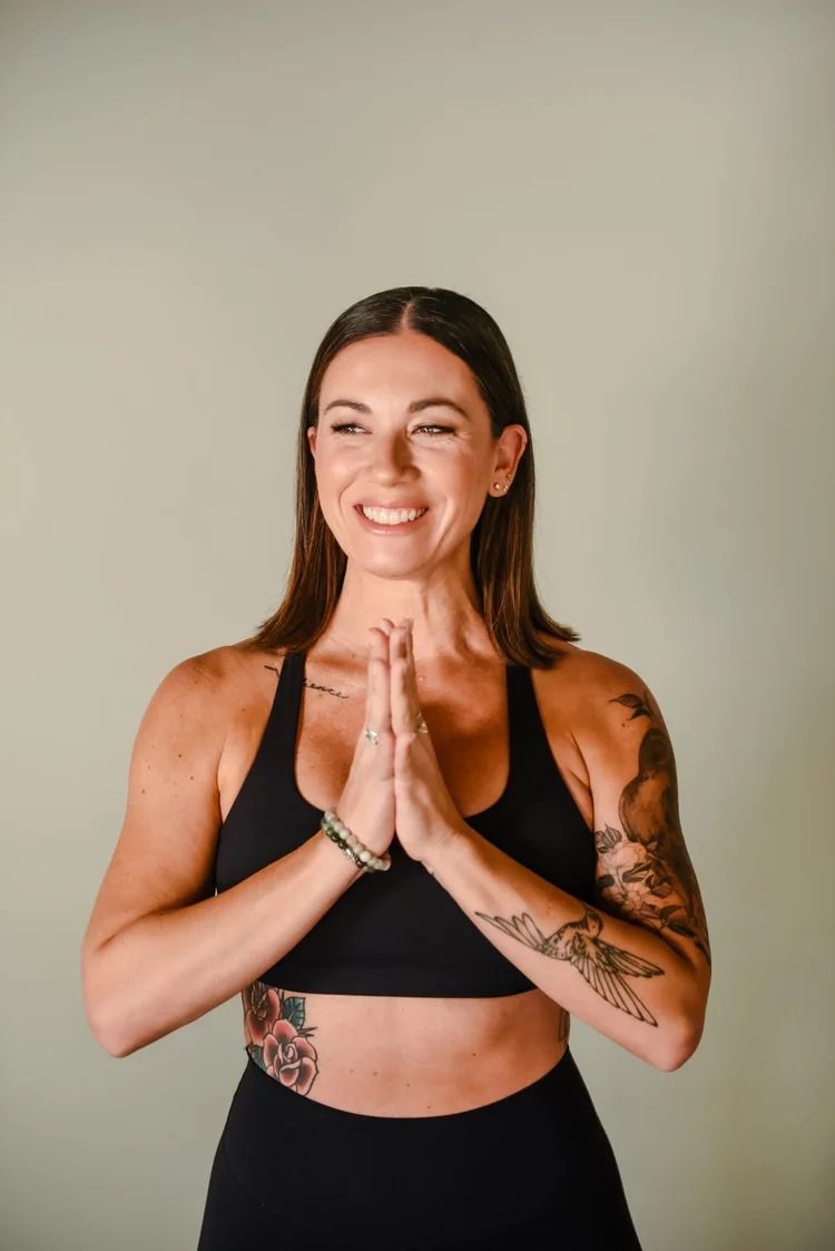 A person in black workout gear with tattoos on their arms and ribs stands with palms pressed together, smiling.