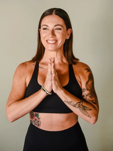 A person in black workout gear with tattoos on their arms and ribs stands with palms pressed together, smiling.