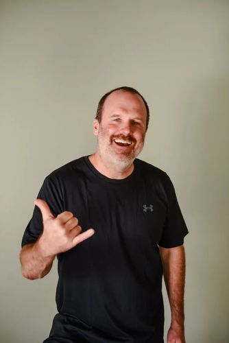 A smiling person wearing a black t-shirt makes a shaka hand gesture against a plain background.