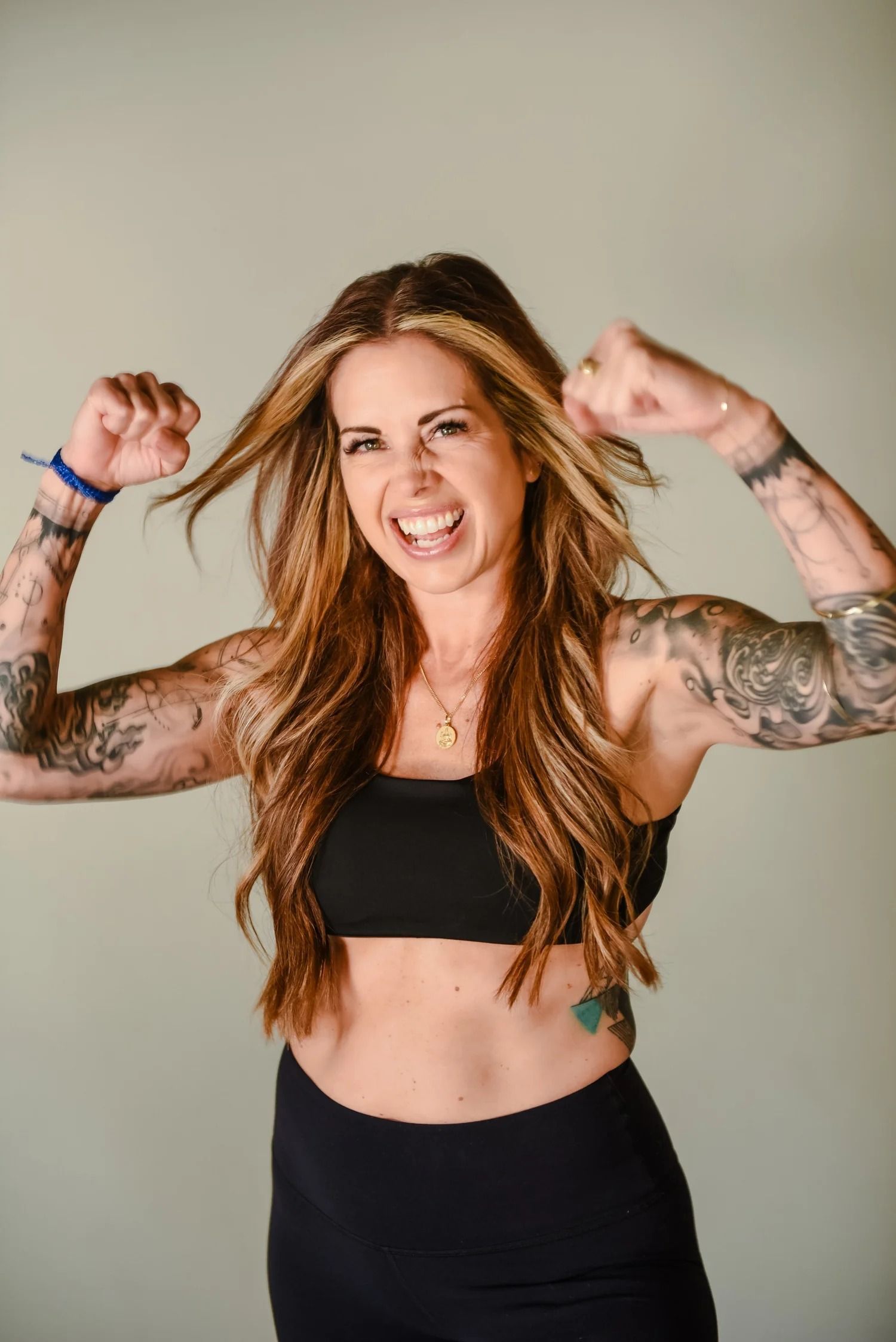 A smiling person with long, crimped hair and full-sleeve tattoos flexes their arms in a celebratory pose.
